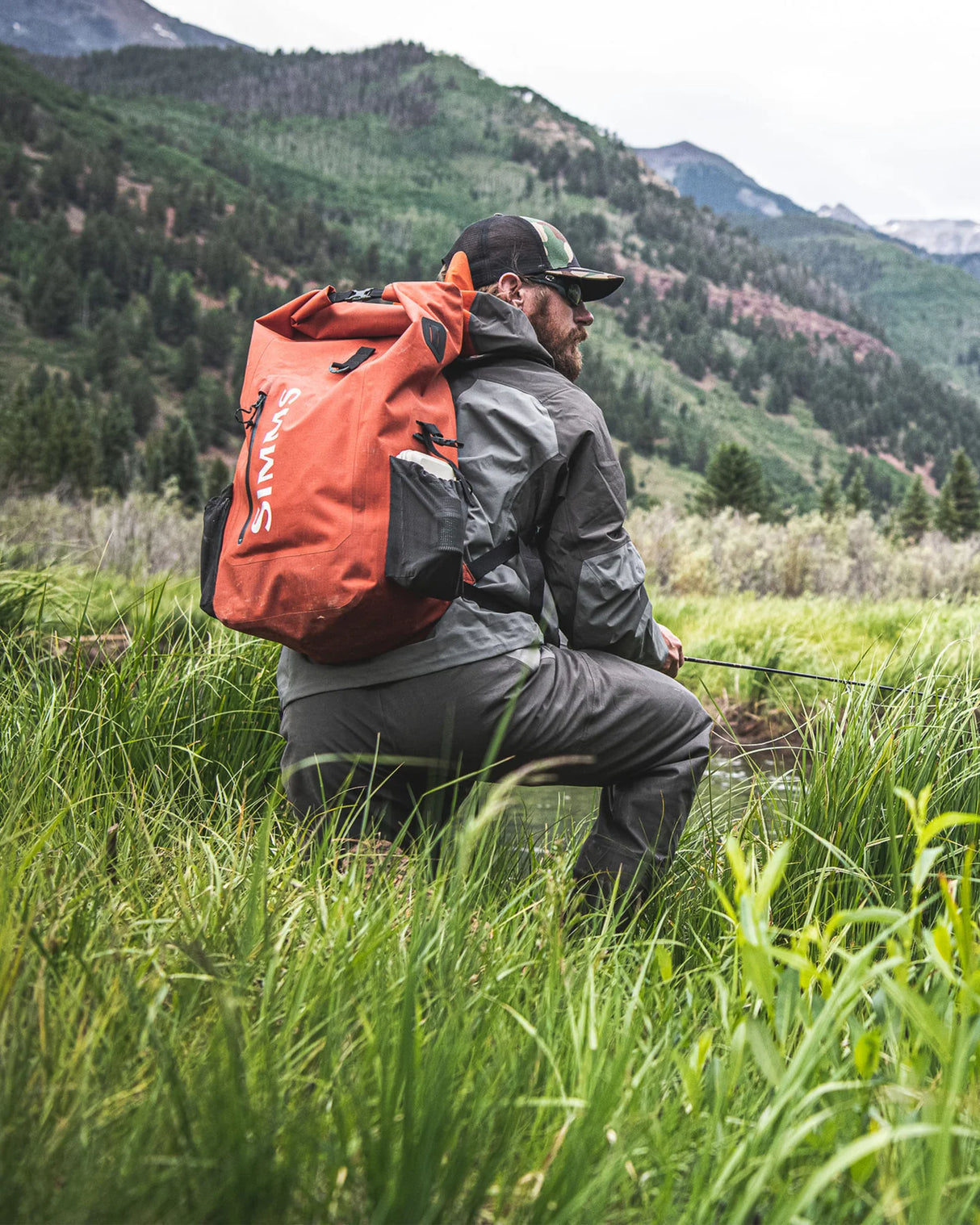 Man with an orange backpack in a grassy field with mountains in the background