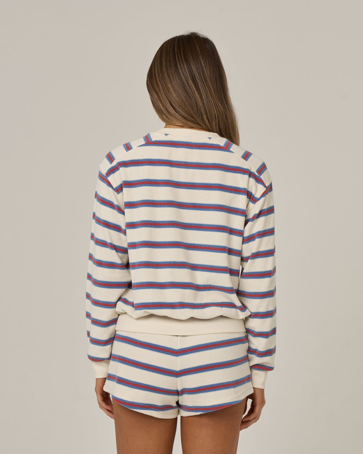 A woman with long brown hair faces away, wearing the Salty Crew Night N Day Crew in off white with matching terry stripe shorts featuring blue, purple, and red lines. The background is plain and neutral.