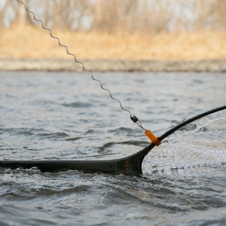 A close-up action shot of a fishing net with a black frame and white mesh, being used in the water. A wire lead line with a small orange connector is attached to the net, set against a blurred background of water and dry grass.