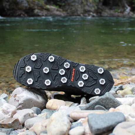 A black outdoor boot sole with metal ice grips resting on a rocky riverbank with a flowing stream in the background.