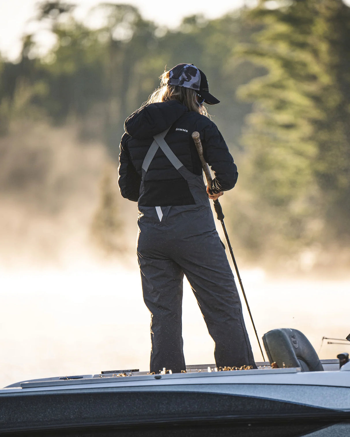 Person standing on a boat wearing a black jacket and cap, holding a fishing rod with a blurred natural background.