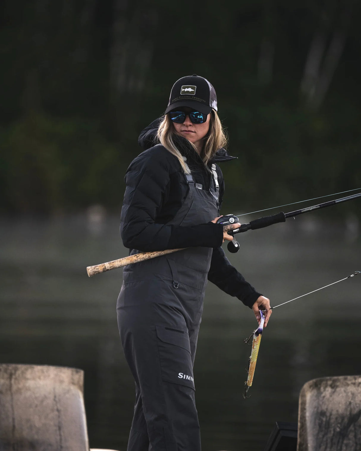 Person in fishing gear holding a fishing rod by a body of water