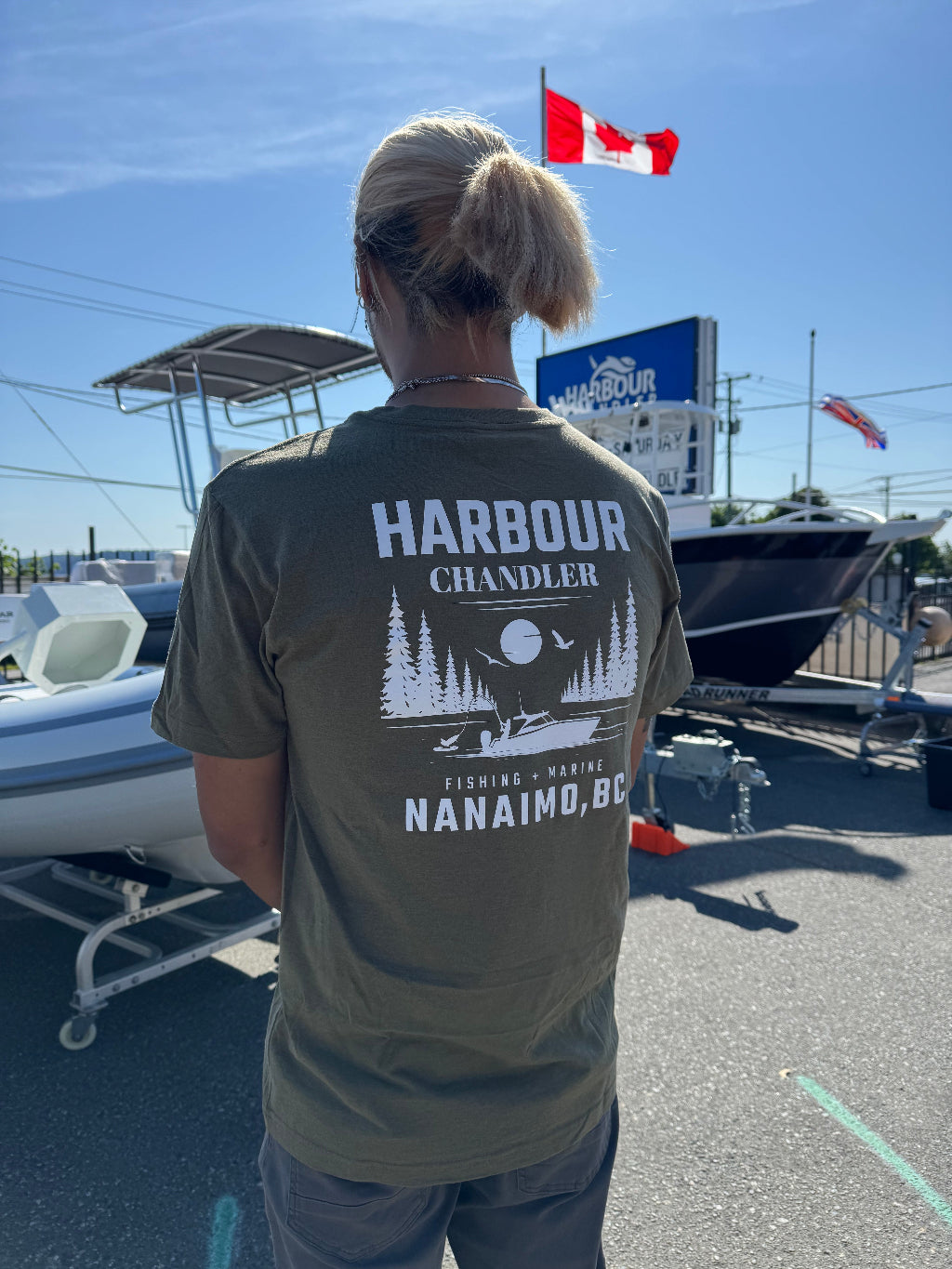 Person wearing a 'Harbour Chandler' t-shirt with a boat and flag in the background - Army Green - L