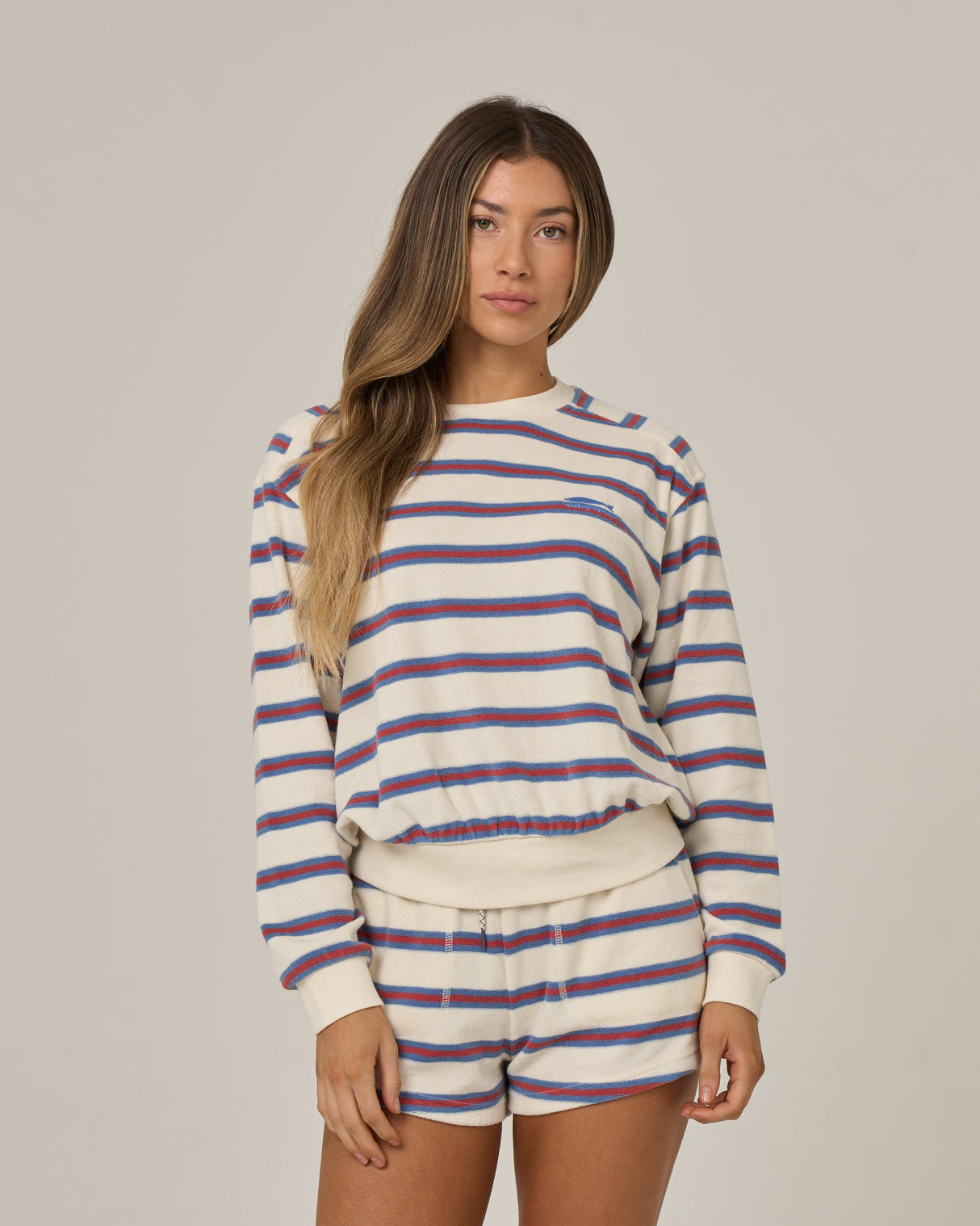 A woman with long, straight hair wears the Salty Crew Night N Day Crew in off white, featuring blue and red stripes, standing against a plain light background.
