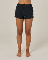 A person stands barefoot on a light surface, wearing Salty Crew Beacons Short - Black, featuring a drawstring waistband and quick-dry fabric, photographed from the waist down against a plain background.
