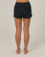 A person stands barefoot facing away from the camera, wearing Salty Crew’s Beacons Short - Black, made from recycled performance fibers with a back right pocket, against a plain light background.