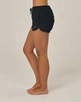 A person stands barefoot on a white background, wearing Salty Crew’s Beacons Short - Black. Made from recycled performance fibers, these shorts feature an elastic waistband, drawstring, and a relaxed fit. Side view shows hands by their sides.