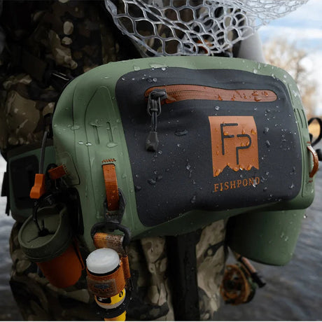 An olive green and black waterproof Fishpond fly-fishing waist bag with copper accents, worn by a fisherman standing in shallow water.