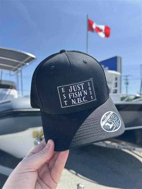 Black cap with text held by a hand against a boat and flag background - Brown with Round Logo
