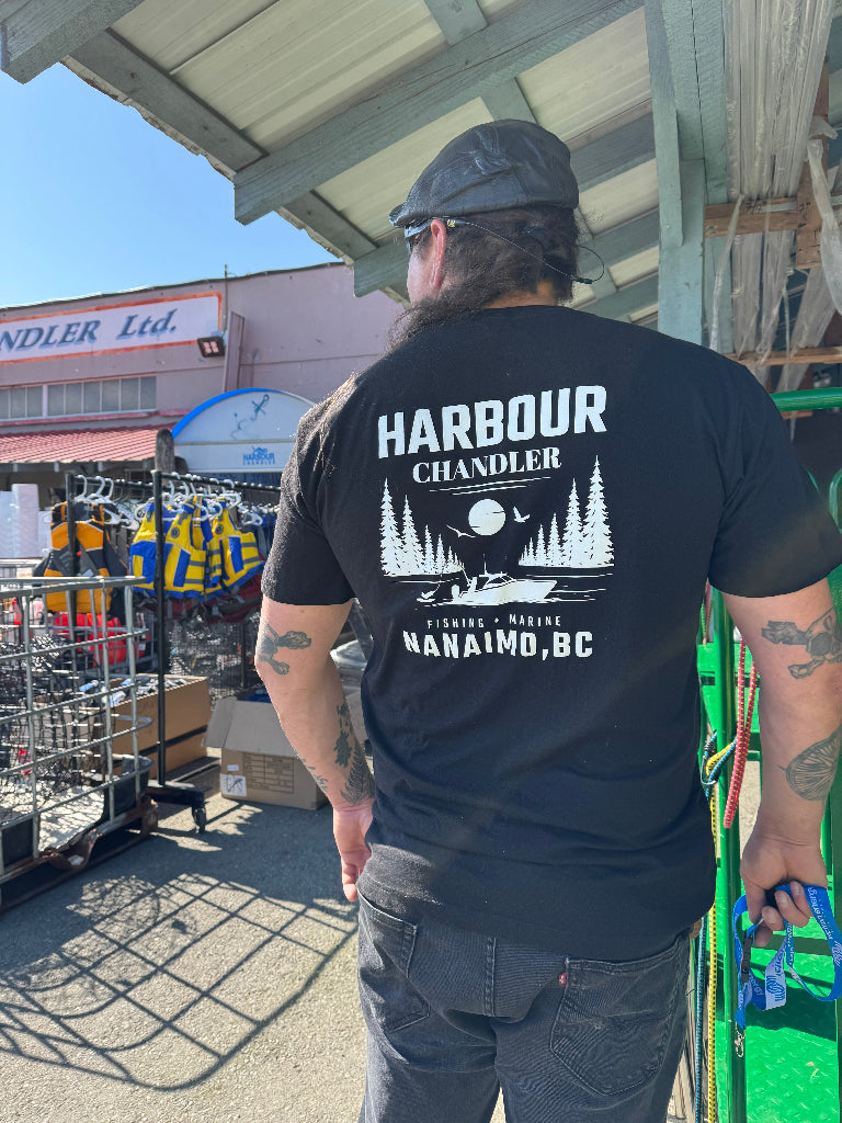 Person wearing a black 'Harbour Chandler' t-shirt with a scenic design, standing outdoors. - Army Green - M