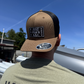 Person wearing a tan and black cap with a visible brand logo, standing outdoors with a clear blue sky.