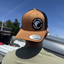 Person wearing a tan and black cap with a logo against a blue sky.