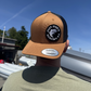 Person wearing a tan and black cap with a logo against a blue sky.