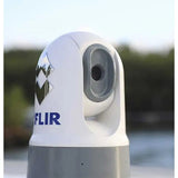 A close-up of a white and gray FLIR thermal camera dome with the blue FLIR logo, set against a blurred outdoor background.