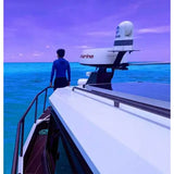 A person in a blue long-sleeve top stands on the deck of a white yacht, looking out over turquoise ocean water under a purple sky. A white marine radar unit is mounted on the yacht's mast.
