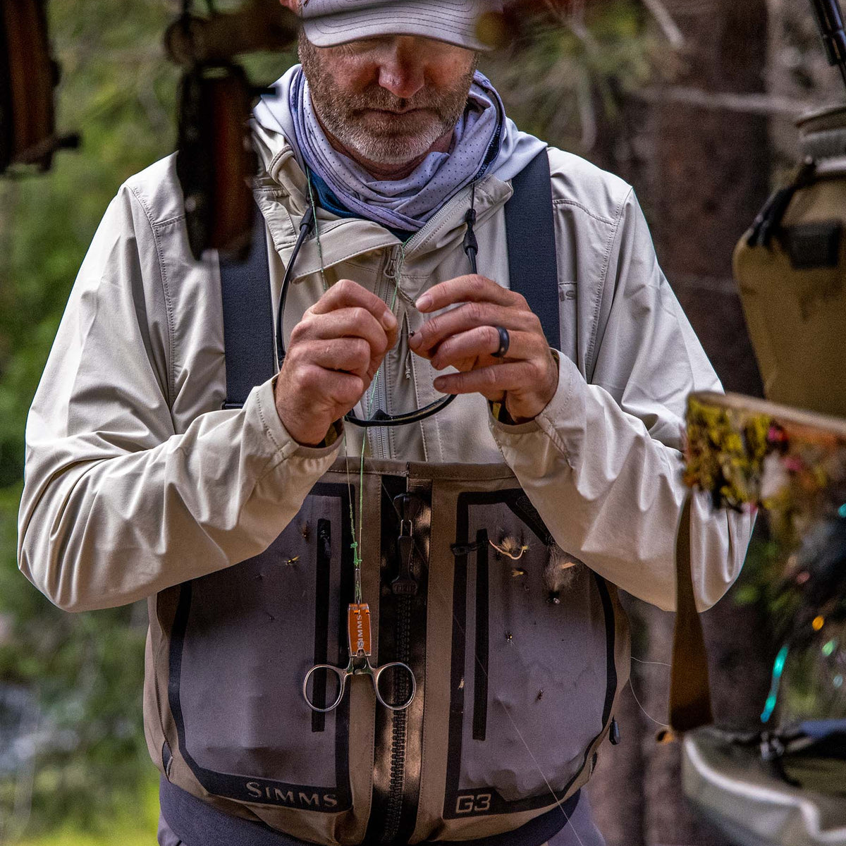 A man wearing a Simms fishing vest and cap outdoors, adjusting a fishing line or hook. The Simms vest is gray and black, and the man wears a beige jacket and a gray bandana.