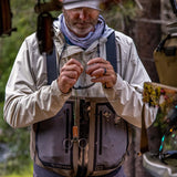 A man wearing a Simms fishing vest and cap outdoors, adjusting a fishing line or hook. The Simms vest is gray and black, and the man wears a beige jacket and a gray bandana.