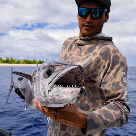A man in sunglasses and a Salty Crew Mariner Lite UV Hood - Tan Camo holds a large, sharp-toothed fish over the water, with a tropical shoreline and partly cloudy sky in the background.