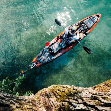 A person paddling a full-sized recreational kayak on clear turquoise water, viewed from an overhead cliffside perspective.