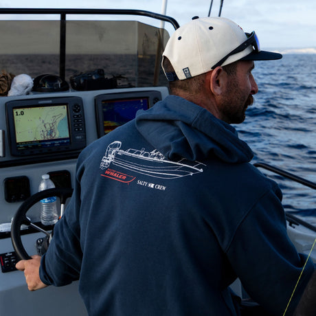 A man steers a boat wearing the Salty Crew Whaler Schematic Fleece in navy, featuring a screenprinted boat graphic and SALTY SIR CREW text. Boat controls, navigation screens, fishing rods, and his white cap are visible.