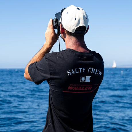 Wearing a black Salty Crew Whaler Icon Crew Tee made from 100% combed ringspun cotton and a white cap, a person stands by the ocean, gazing through binoculars on a sunny day as a sailboat appears in the distance.